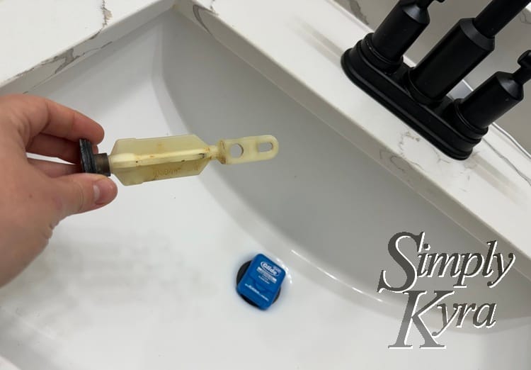 Image shows a blue container of dental floss over the sink drain while I hold a clean stopper up in front. 