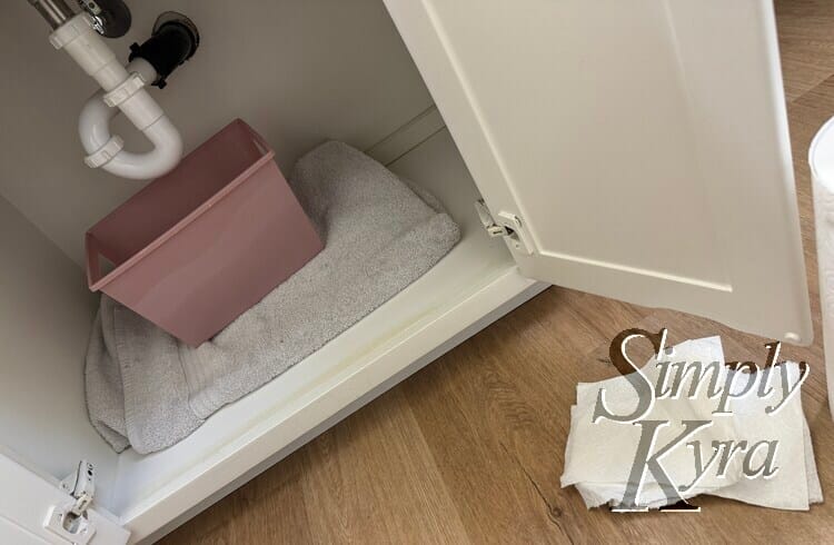Image shows under my sink with a plastic bucket ready to catch water, a towel, and some paper towels nearby. 