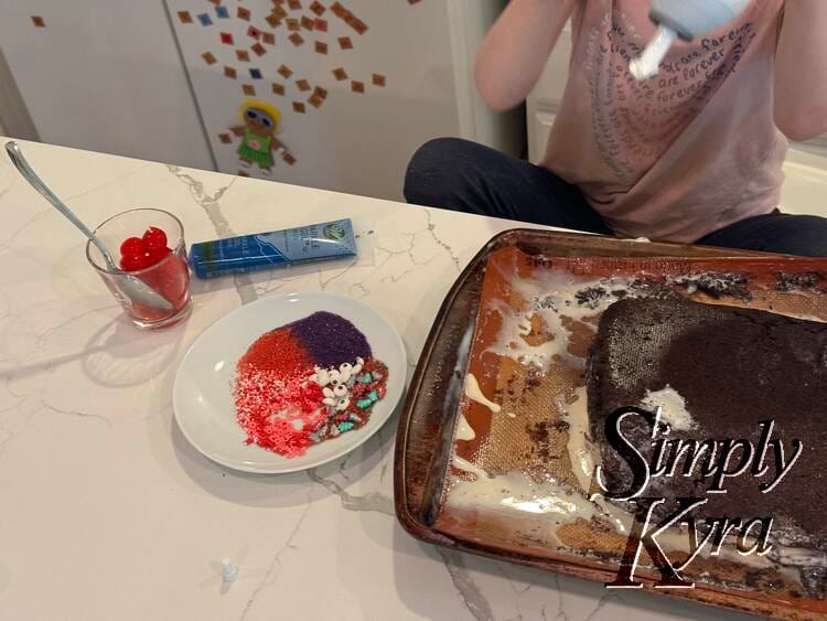 The messy melting but now brownie covered cake is centered on the silpat lined cookie sheet with whipped cream above and laid on icing writer, cherries, and sprinkles of Zoey's choosing on the left.