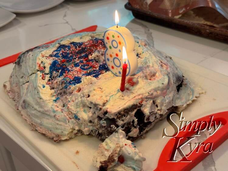 A corner closest is broken off but the cake is laid out on a red edged white cutting board and the cake has two candles light up.