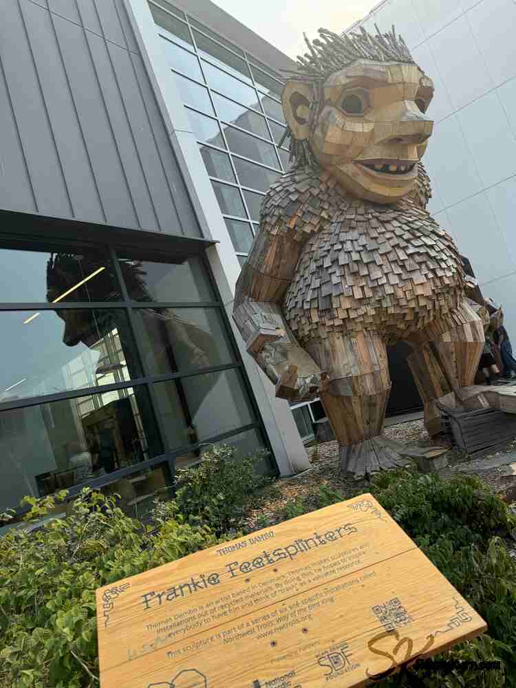 The sign is tucked in the plant while es stands before you in front of a building. Less nature and of his Troll-like. 