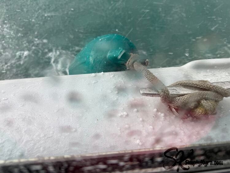 Image shows the rain splatted window and, through it, the side of the boat with hail sitting on the edge. 