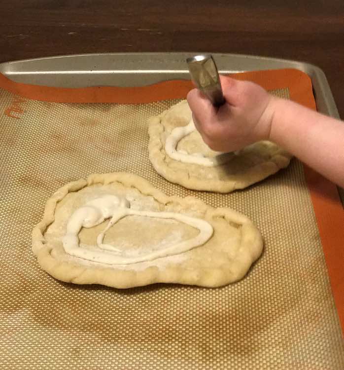 Closeup of Ada spreading ranch dressing on her pizza crust with a spoon. Underneath the two pizza crusts are a silpat and cookie sheet.