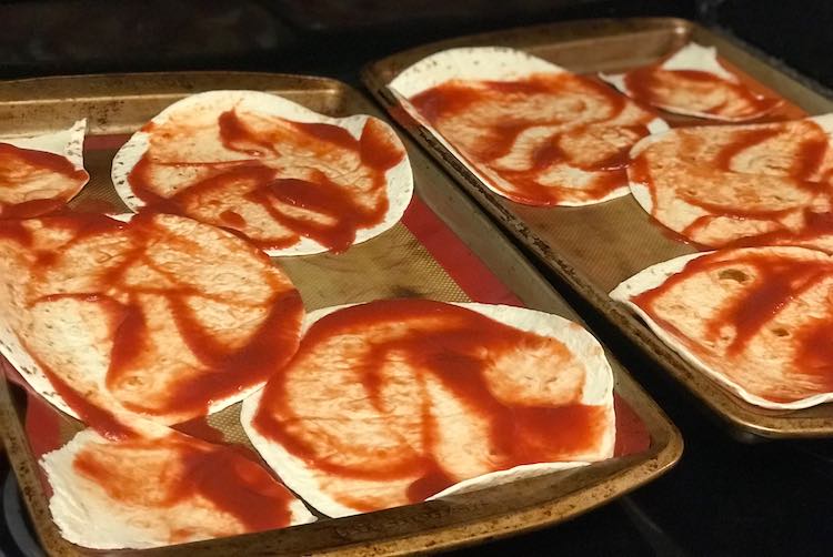 Closeup of the two cookie sheets with the tomato sauce covered flour tortillas.