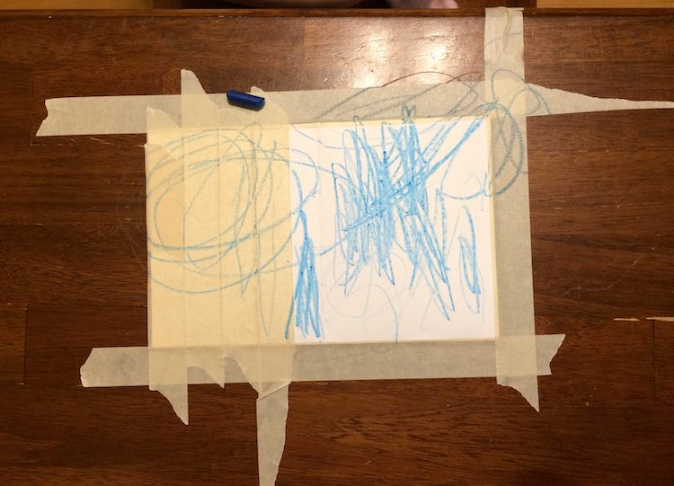 Taped down card on a table showing a square of white. There's a blue crayon laying on the card and blue scribbles over the entire thing.