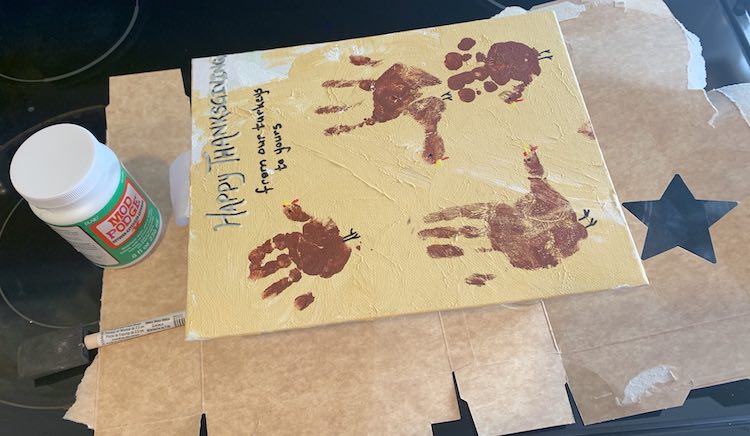 The Thanksgiving turkey handprint canvas is shown below the camera. The glasses are hidden below the canvas and the cardboard is sticking out from underneath. The bottle of mod podge and the foam brush are to the left of the canvas.