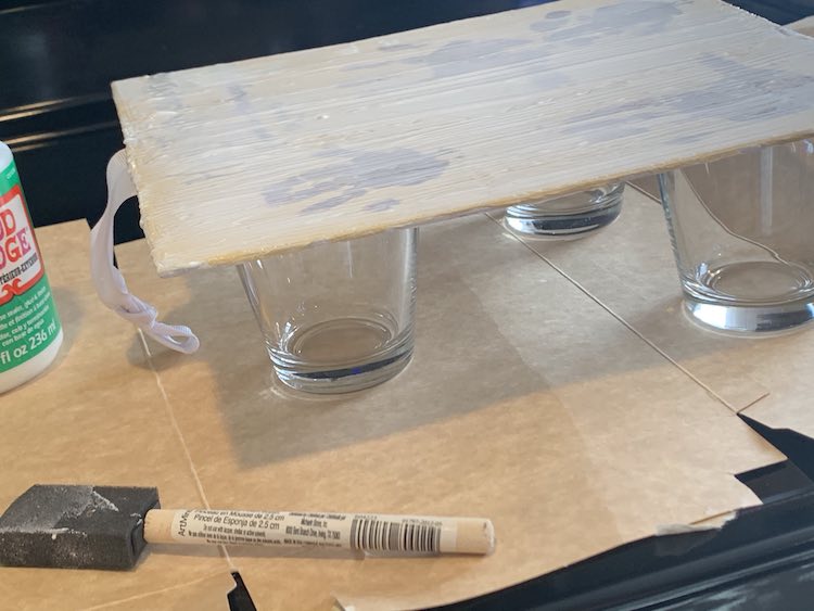 Side view of the canvas allows you to see the drinking glasses holding the canvas up. There are two glasses at either of the bottom corners and one centered at the top. The ribbon at the top of the canvas dangles down and brushes the cardboard. The used foam brush and closed mod podge bottle are visible at the bottom side.