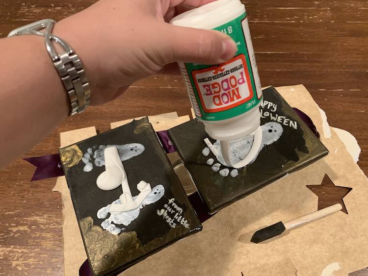 Both canvases laid out suspended over the cardboard below. The bottom canvas already has mod podge laid out on it and the top one is currently getting it poured onto it.