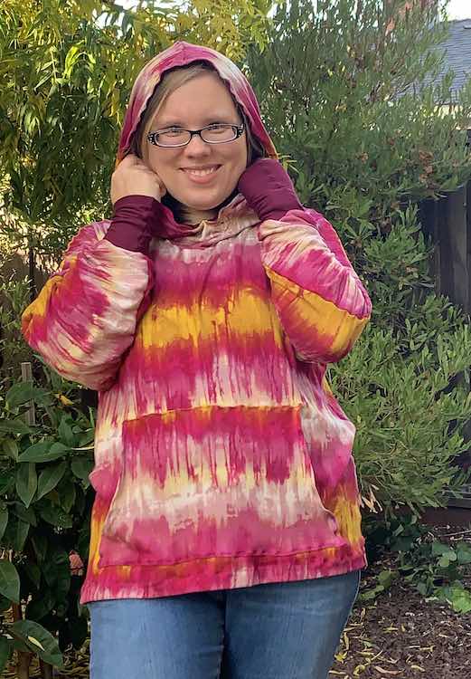 Front view of the hoodie with the hood on over my head. My arms are upright holding onto the hood which better shows off the cuff at the end of the sleeves.