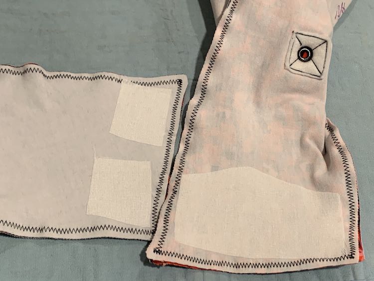 Image shows the interfaced flaps of both kids. On the left is the horizontal laying flap showing two smaller squares of interfacing. The right side shows a vertical laying flap with a large rectangular-ish piece of interfacing. You can also see the underside of the grommet near the top of the image.