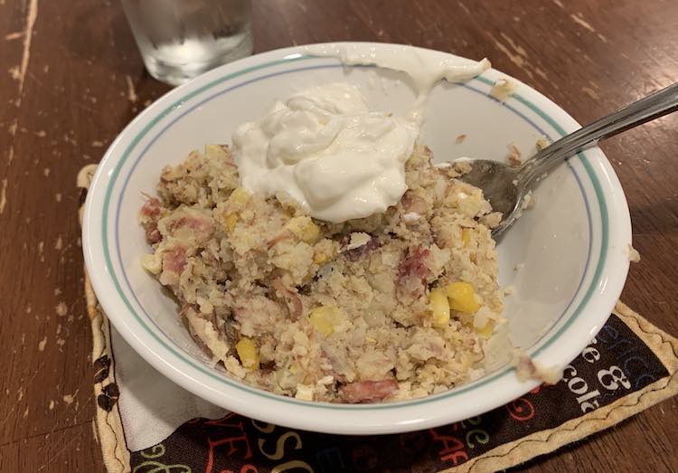 White bowl with blue encircling the bowl near the top. Contained inside of it is another type of mashed potato perogies dish this time with corn and cooked in a pot. It is topped with a dollop of Greek yogurt.