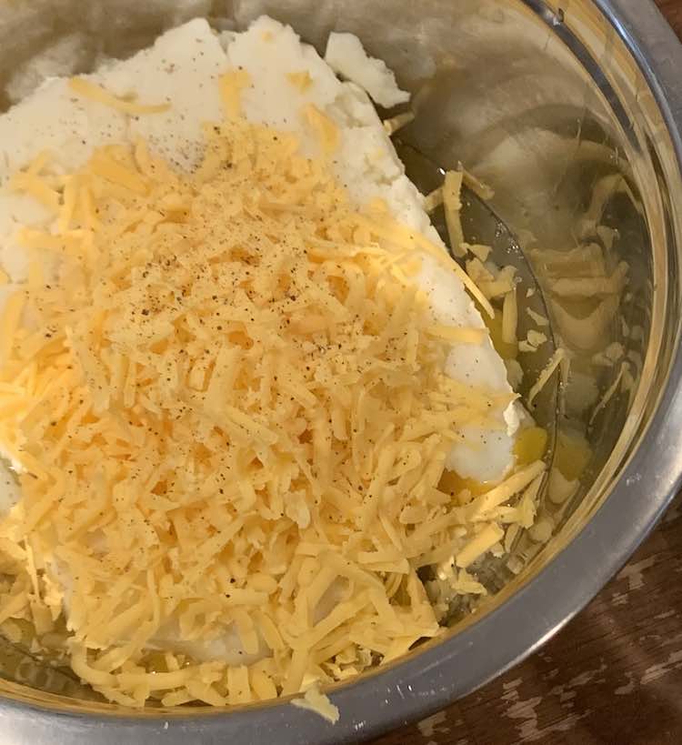 Metal bowl containing mashed potatoes with grated orange cheese on to and then black flecks of pepper on top of that.