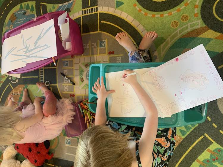 Image shows an overview of Ada and Zoey sitting on the car mat drawing on lap desks. Zoey is using decorative washi tape to attach to pieces of paper together artistically while Ada, to the right, follows the video to draw the pigeon from Mo Willems' book.