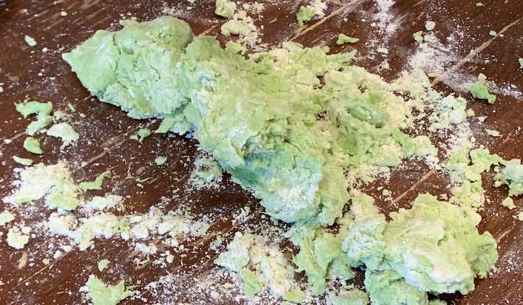 Clumps of green playdough coated, on parts with white. Around the clumps, resting on the table, are lots of white powder from the flour that wasn't able to be mixed in.