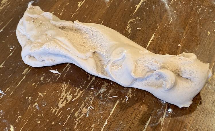 The light brown playdough is resting on a dark brown table. The playdough has been pulled into a cylinder shape though the grooves are diagonally across the playdough rather than horizontally straight across like before.