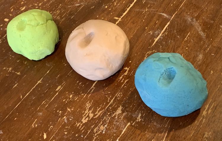 Three balls of playdough rest of the table all with a finger mark on the top to show how the playdough squished in. The green is furthest away, to the left, with the blue closest, to the right. The brown is resting in the middle.