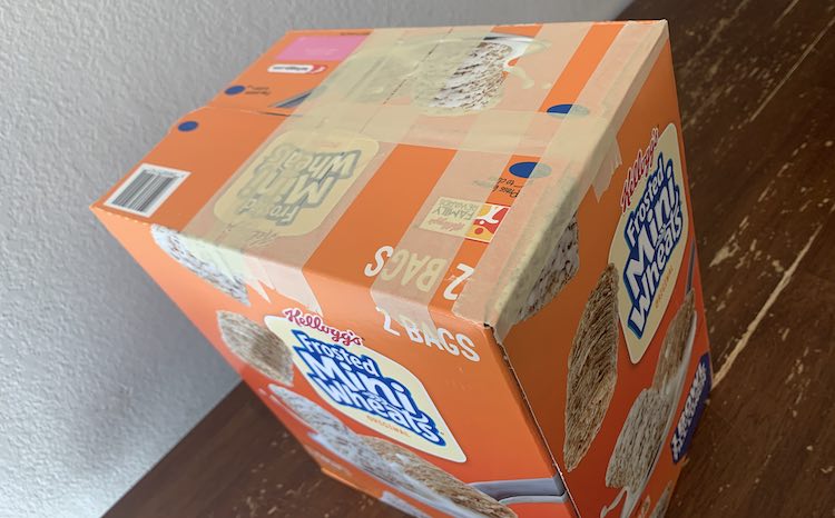View, angled, of the top of the box (Frosted Mini Wheats) with beige masking tape holding down the top flaps in the center and on the one corner edge.