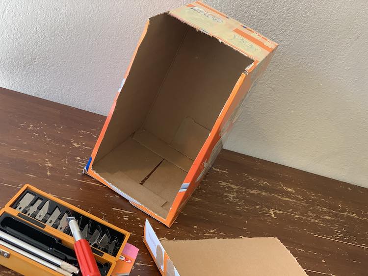 View of the box missing most of it's front and some of it's top so you can see the brown inside of it. In the foreground, at the bottom of the image, you can see the opened X-Acto box, the knife, the blades, and the removed part of the box all going off photo.