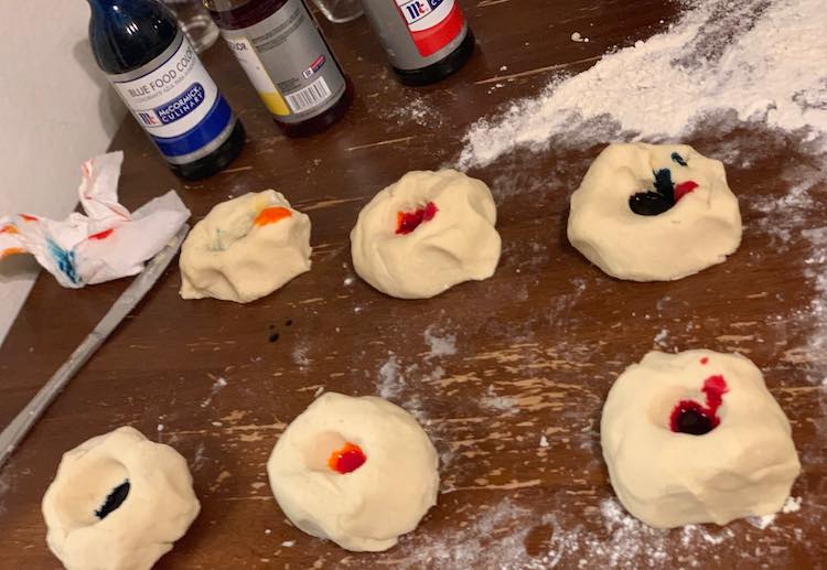 Image shows six mounds of white playdough with divots in the center. They each have different drops of red, blue, or yellow food dye in the center. The large bottles sit in the back beside a scattering of white flour and a splotched paper towel. 
