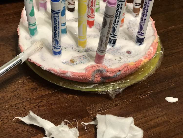 Closeup of the base of the felt pen holder. You can see the glitter and a couple of gemstones where the white mod podge was scraped away. The paintbrush used rests leaned up on it at the left while the excess wipes are at the bottom of the photo.