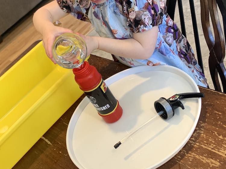 Closeup of Zoey pouring water into the opened fire extinguisher toy standing on a plastic tray. The lid lays beside it and the yellow sensory bin lays behind it.