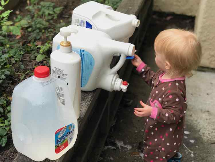Zoey is shown attempting to get water out of the jug by holding up a mould against the spigot without trying to turn it on. Lined up next to the bottle is another detergent bottle, shampoo pump bottle, and a filled milk jug lined up going towards you.