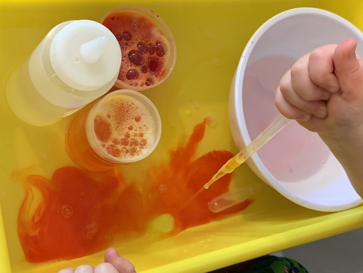 Again an overview of the yellow bin with a white bowl with red tinged water in the base. To the left is a squeeze bottle with clear liquid and two cups filled with yellow and red dye. The part of the bin at the bottom of the image shows Zoey forcing yellow dye out of her pipette causing an expanding orange cloud permeating through the clear water from that spot.