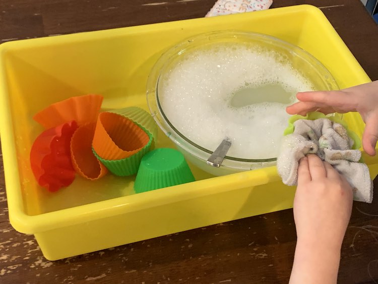 Partially overview of the yellow bin. The right side holds a glass bowl filled with soapy water and a spoon. Over the bowl Zoey's hands are washing a yellow cupcake liner with a small washcloth. To the left, in the yellow bin, a pile of cupcake liners sit waiting to be washed.