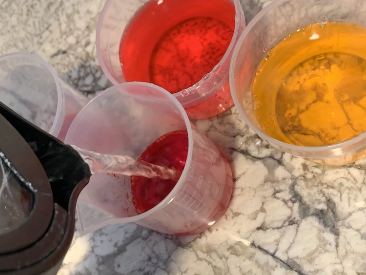 Closeup of the containers of food dyed water. The, slightly hidden, one on the far left only has dye while the two back ones have water added already. The bottom-most one is getting filled with room-temperature water from the kettle as it seemed