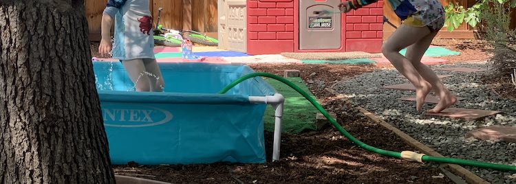 Short and wide image showing the Intex pool (left side of image with a sprinkler spraying up a bit and Zoey in it. To the right you can see the garden hose coming out over the side of the pool and Ada's feet as she rushes over.
