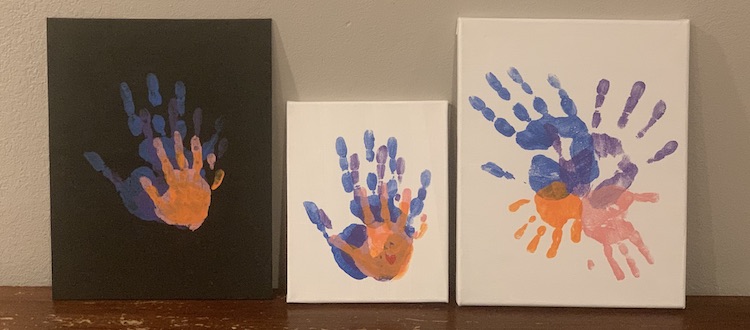 The three canvases leaning against the wall and sitting on the table. They're placed beside it each other so from left to right we have:
1. the black canvas with the handprints stacked overtop of each other from biggest to smallest.
2. The smallest canvas with the white background. The hands are the same color and are stacked like the left one. In the lower right corner of the palm there sits a red heart.
3. The white large canvas with the handprints forming an X with the largest handprints forming the top and the smallest ones forming the bottom legs of the X.