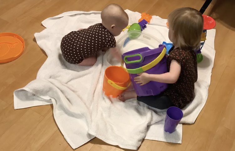 Zoey's hand reaches into the water as Ada (toddler) lifts the large bucket enough to pour more water into the plastic container.