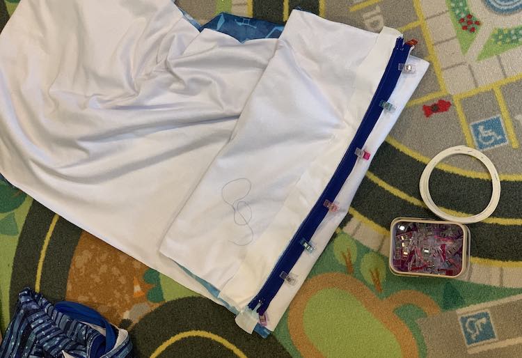 Image is captured looking down at the pillow cover from above. It's laid out on my car mat with a container of sewing clips and wash-away tape next to it. The fabric was folded right sides together with the zipper zipped closed. The zipper end of the cover is folded over a couple times but still shows the zipper with the sewing clips attached to the non-sewn side.