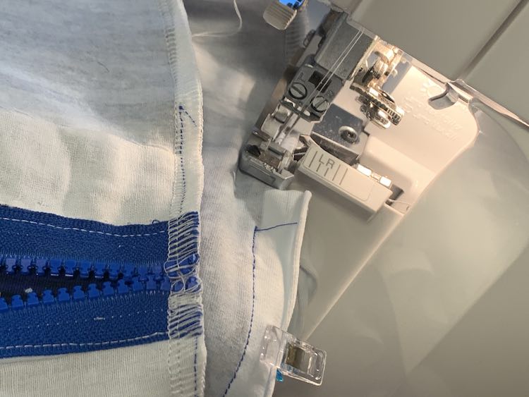 Closeup of the serger while closing the side of the pillow. The needle has been paused to take the photo but the pressure foot is about to reach the sewing machine's stitching so it's a couple inches from the zipper tape. The other end of the zipper has been folded over so you can see where the serger went over the zipper end and sewing machine's stitches and how it looks a bit looser there.