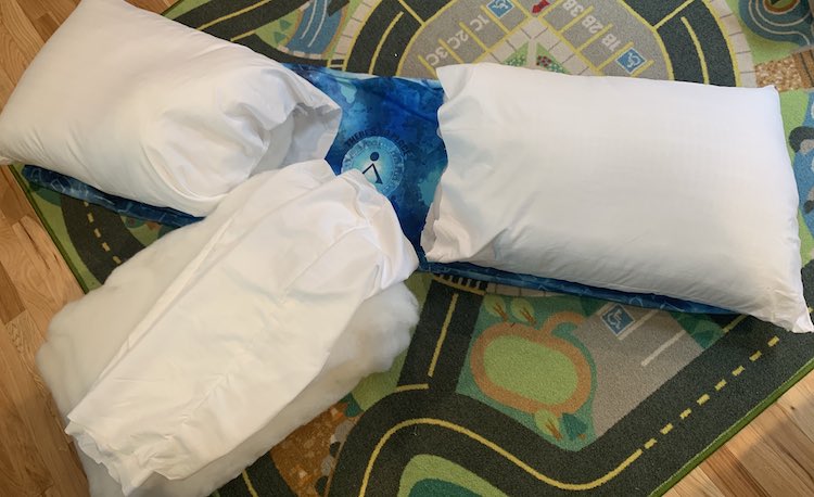 Image shows the long pillow cover laid out on the car mat mostly covered by the two pillows with the one end open. The center panel shows through but covering the bottom of it, angled up, lays the stuffing of the third pillow with it's pillow covering flopped overtop.