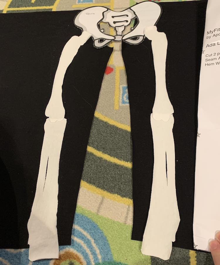 Image shows two black flat fabric legs laid out with the matching crotch lines facing at the top. You can see part of the paper pattern piece being held over the half of the right leg. At the top center is the paper pelvic bone and descending from there are the upper and lower leg bones. The bottom of the leg bone stretches pasts the bottom of the pant leg by a bit. 