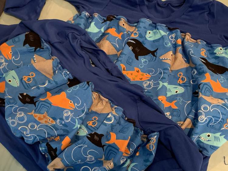 Image shows two shirts laid partially on top of each other on the ironing mat. Both garments have dark blue knit at the top and bottom with blue shark filled fabric in the center. The smaller shirt has one grey shark with orange hearts drawn on it while the back shirt has an orange shark with two hues of blue hearts on it.