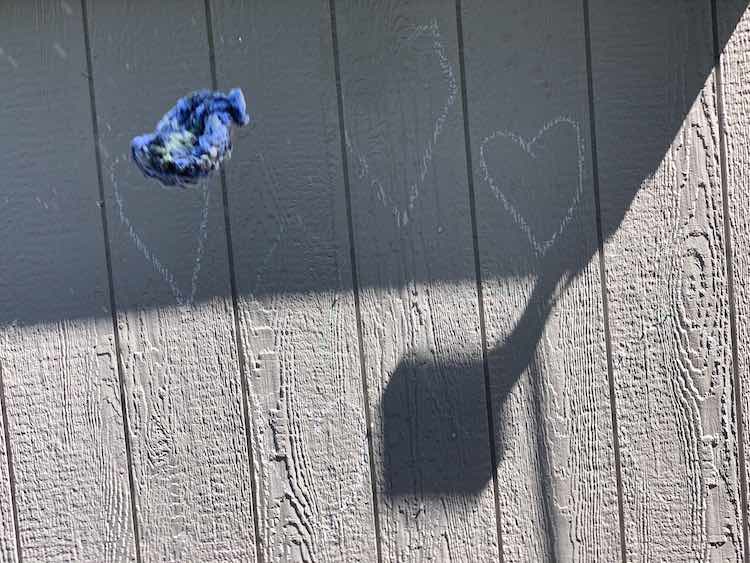 Image shows a shadow covered shed with some white outlined shapes drawn onto the wall. In the foreground is a single wet yarn water balloon flying towards the wall and spraying water out the back (above). 