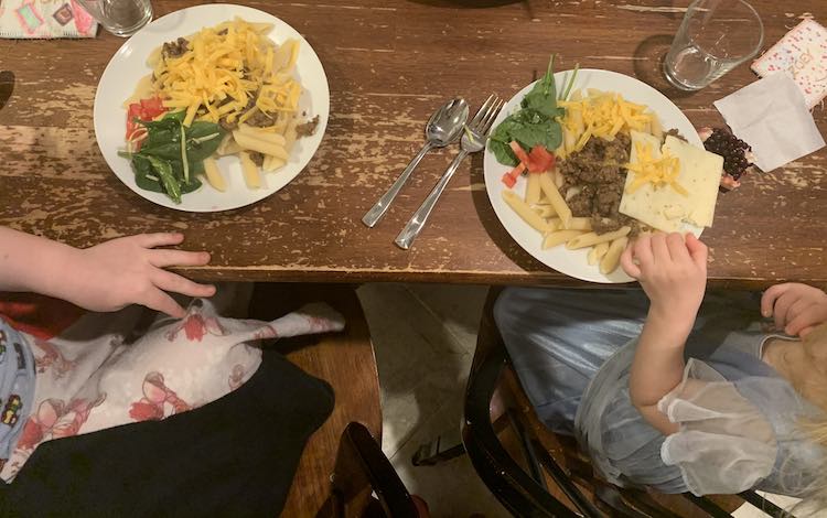 Image shows both kids sitting at the kitchen table with a plate of penne pasta, taco meat, melting cheese, spinach, and diced tomatoes on top.