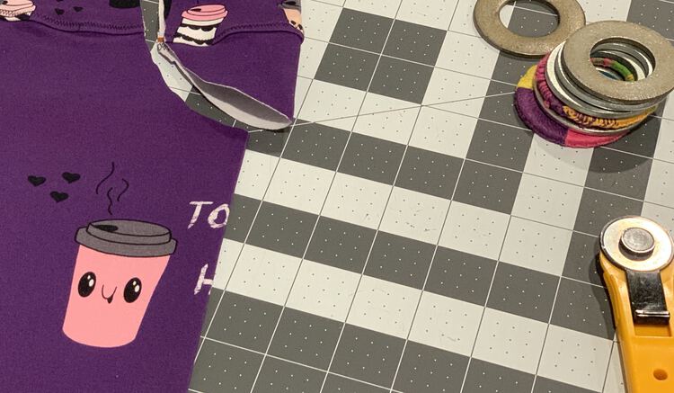 Image shows a closeup of the folded over fabric with an adorable lidded cup on it. Part of the fabric is cut leading past a topstitched seam where the panel was extended into a patterned fabric. To the right, still on the cutting mat, sits a rotary cutter and several pattern weights.