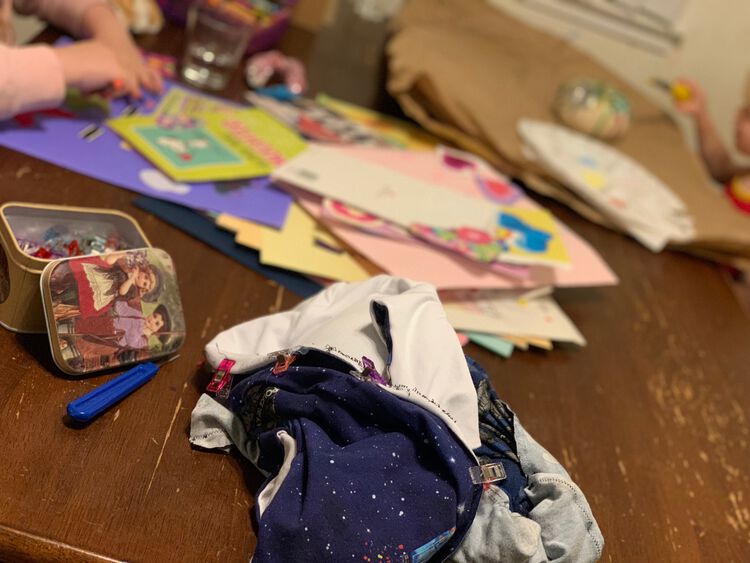 Image shows the dress bunched up on the table with sewing clips attached and black thread sewing. The seam ripper and container of sewing clips sits beside it. In the back and slightly blurred you can see one kid painting a white pumpkin and another cutting up leftover greeting cards.