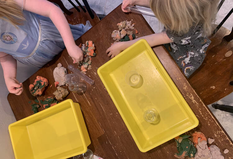 Image shows the kids set up at the end of the table. Both have a yellow bin in front of the and are planning their volcano.