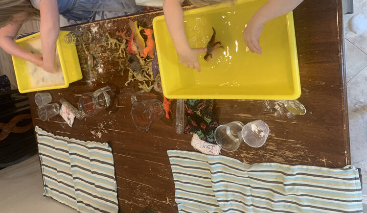 Image shows the entire table. Both girls have their hands in their bin with the dirty dinosaurs between them. The other side of the table has a clean tea towel laid out while between the two is their drinking glass (got dumped and a clean given), name tag, and extra tools.