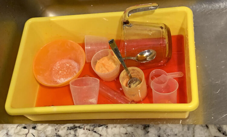 Image shows a yellow bin with orange-themed items inside it. The bin is sitting in the metal sink with some of the counter showing at the bottom of the image. The bin includes two plastic cups with orange tinted wet baking soda, several empty plastic vials and cups, two spoons, an white powder dirtied orange bowl, and a measuring jug on it's side.