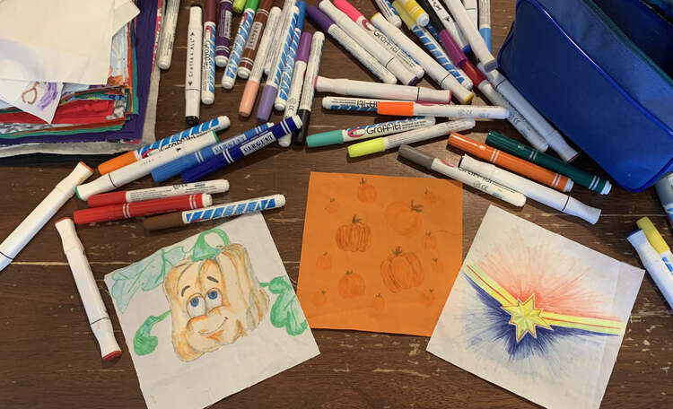 Image shows the final three decorated squares of fabric that I drew on for the girls. In the center is an orange square with pumpkins while to the left there's a spookley on a white square and to the right a Captain Marvel logo. Above it lays an empty blue pencil pouch, a stack of the remaining masks, and lots of fabric markers.