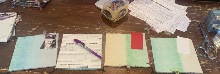 Image shows a stack of masks to the left and a stack of patterns in the upper right. In the main, bottom, of the photo sits four squares of fabric. The one second from the left has a pattern piece on it and a pen that just marked the opening at the top and bottom. The other three squares are already marked and have the extra two rectangles stacked at the top. Some are clipped together and some are still waiting for the clips.