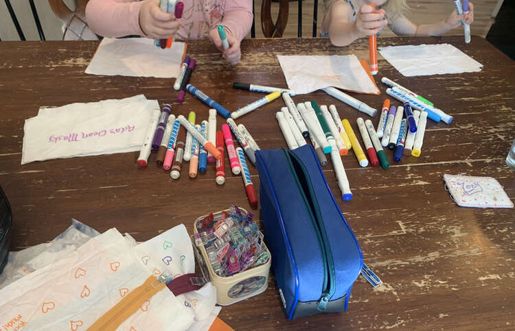 Image shows the kids grabbing fabric markers from a pile in the center while white rectangles of fabric sit in front of them. Closer to the bottom sits a pile of clips, and empty pouch, and a stack of fabric and zippers.