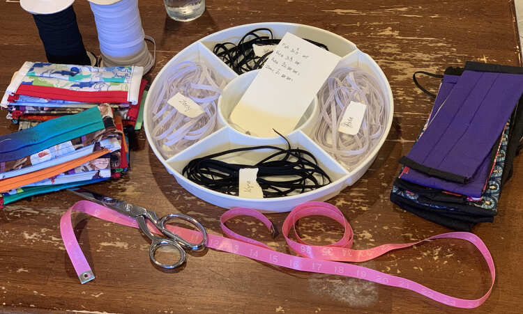 Image shows the measuring tape and scissors I used to cut the elastic at the bottom of the image. Above it sits a five sectioned container base with four piles of elastic and a paper with a name placed on top. To the sides sits piles of finished masks and two rolls of quarter inch elastic in black (for the adults) and white (for the kids).