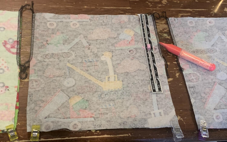 Image shows the back square of fabric with two clips at the bottom, black serged edge on the top 3.5 inches on either side, a ruler on the right, and a mark at the 2 inch spot. To the left and right you can see the edge of another one.