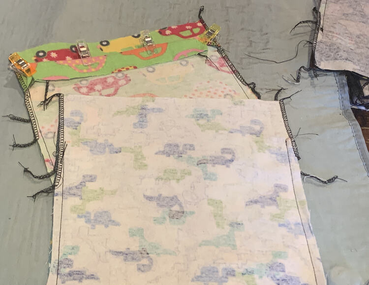 Image shows two bags overlapping each other with a stack of several off photo to the right. The top bag is inside out and sewn up to the top two inches. The bag underneath has the front side folded down with clips holding it in place.